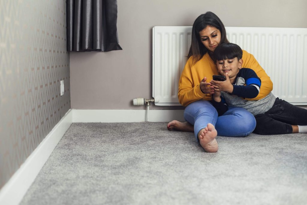 Woman sitting on the floor with a young child in front of a radiator looking at a phone.